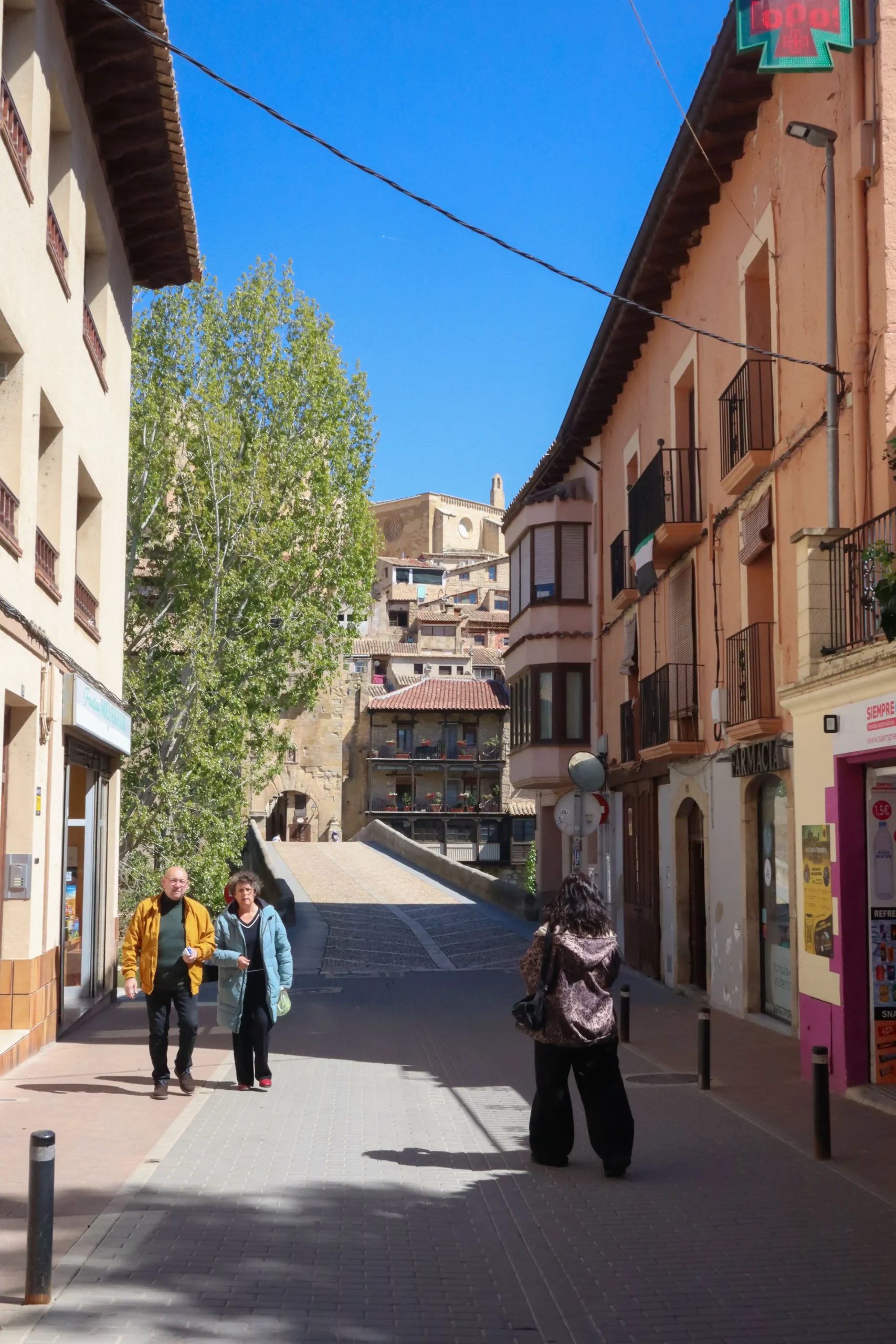 Calle peatonal con personas caminando hacia el casco histórico de Valderrobres con edificios tradicionales y el puente de acceso al fondo