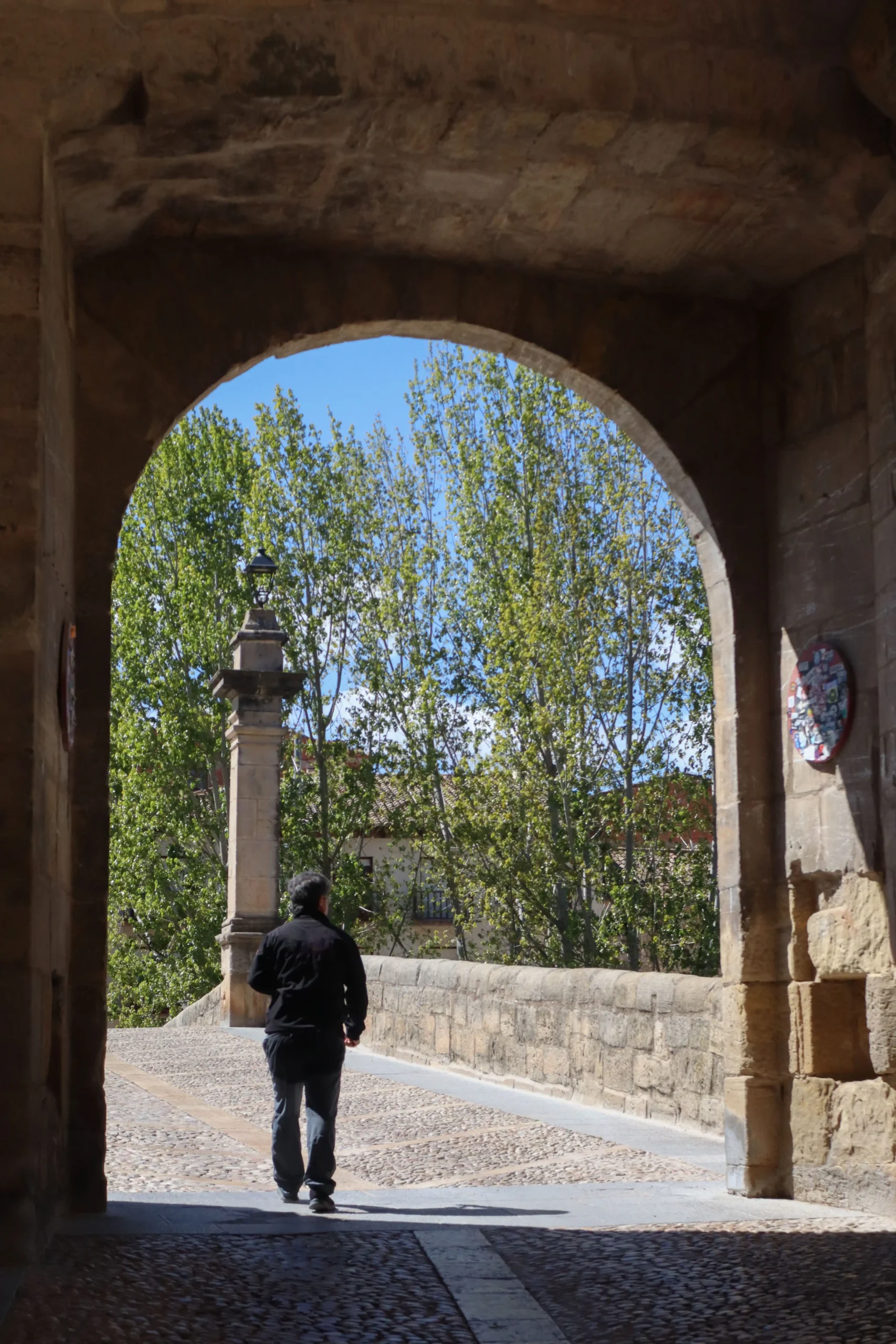 Persona caminando bajo el arco del Portal de San Roque hacia el puente de piedra de Valderrobres rodeado de árboles
